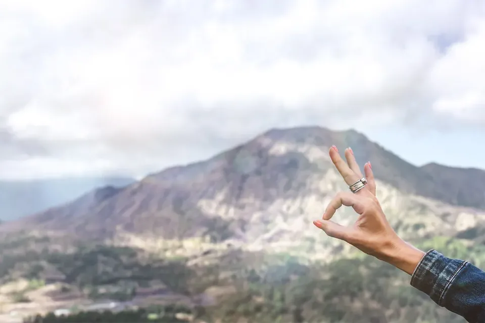 A hand making the OK sign in front of a mountain under a cloudy sky
