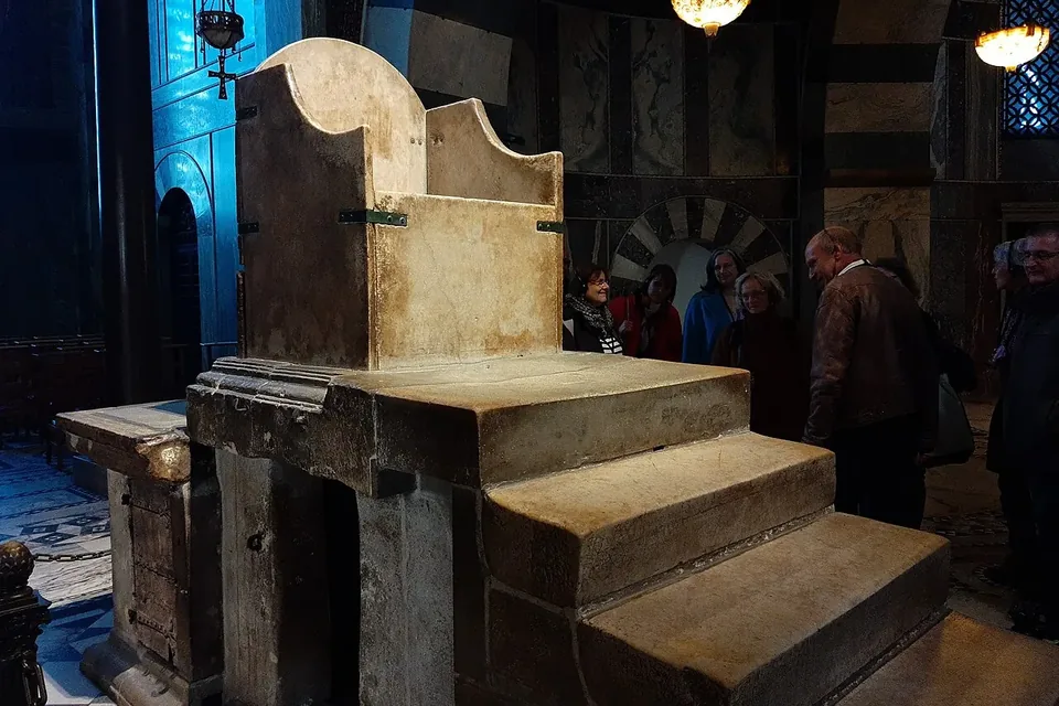 A plain stone throne at the top of six stone steps inside a dimly lit cathedral gallery