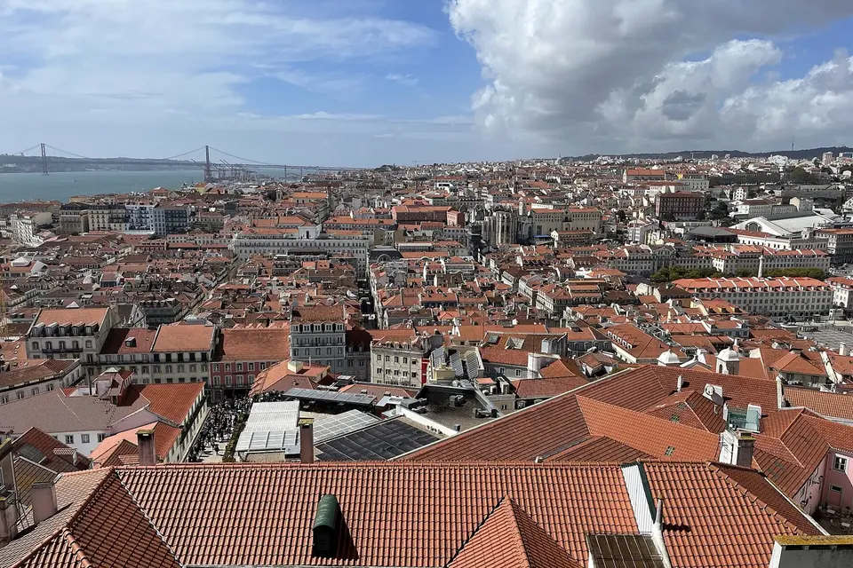 Terracotta rooftops of central Lisbon stretching to the river, with a red suspension bridge in the distance under a cloudy sky